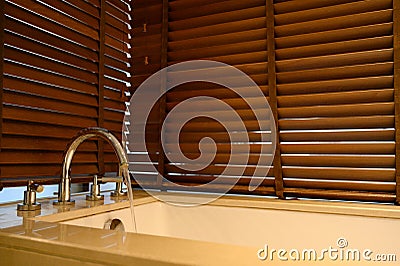 Close up ofÂ water flow from water tap into bathtub with wooden venetian blinds on background in bathroom Stock Photo